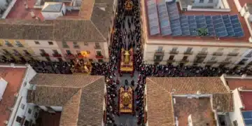Vista aérea de una procesión de Semana Santa en Sevilla con pasos ornamentados en calles adoquinadas, contrastando con paneles solares modernos en tejados, un dron verde en el cielo, y una mezcla de luz cálida y fría.